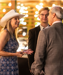 11.15.25 - Party - Finals - I-124 A woman in a white cowboy hat laughs while chatting with a group at an evening outdoor party, string lights glowing in the background.