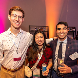 resident fellow pm25 Three smiling colleagues pose together at an indoor event with colorful stage lighting in the background.
