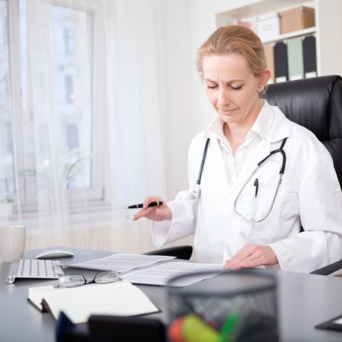 A female doctor in white coat with stethoscope, seated at a desk, reading printed paperwork. Coffee cup, computer keyboard, and reading glasses also on desk. The doctor is seated near a window with a white sheer curtain.