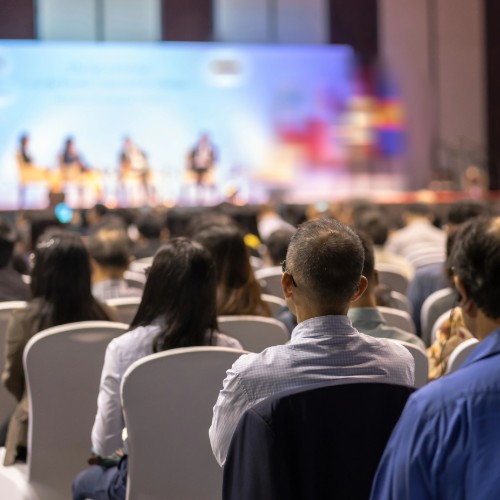 Attendees in theater-style seating watching a presentation by multiple speakers